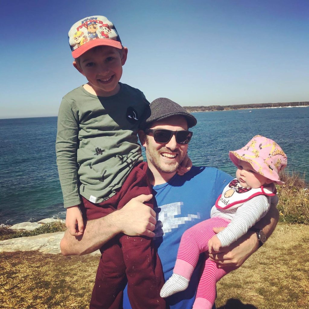 Joel Scully smiling by the ocean, holding his son Cassius on his shoulder and his baby daughter Juniper